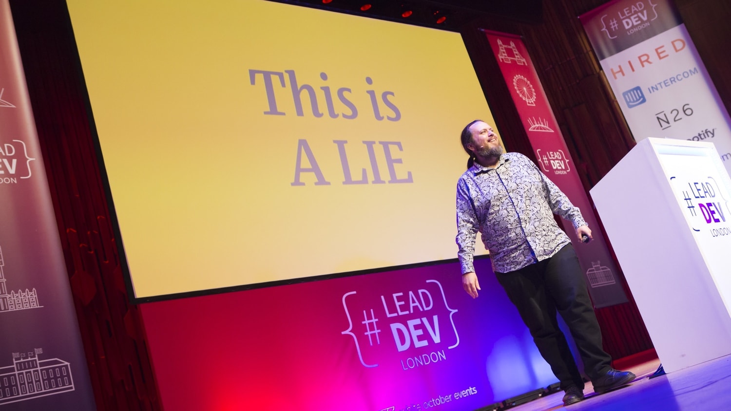 A somewhat pretentious picture of Adrian giving a talk at Lead Developer London 2018. Look at him. Grinning like a loon. For shame. There's a big slide projected behind him reading 'This is a LIE'